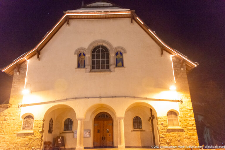 Illuminated gabled facade of St. Mauritius Church with three-arched porch and religious figures beside the upper window.
