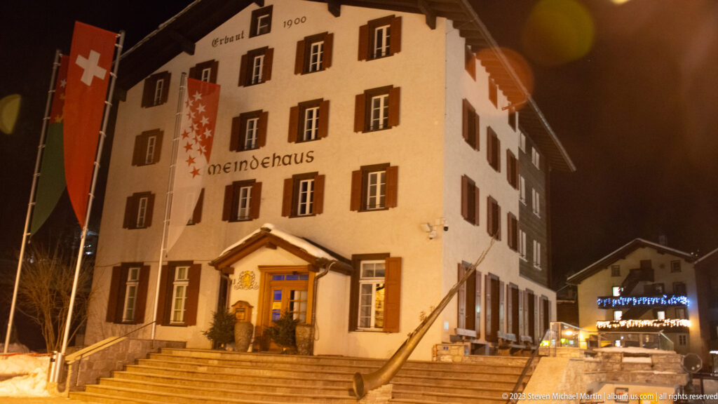 Illuminated white Gemeindehaus with dark shutters, Valais emblem, flags, and broad steps at night.