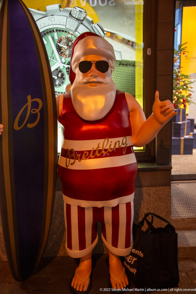 Santa figure in sunglasses and striped trousers beside an oversized Breitling display outside a shop.