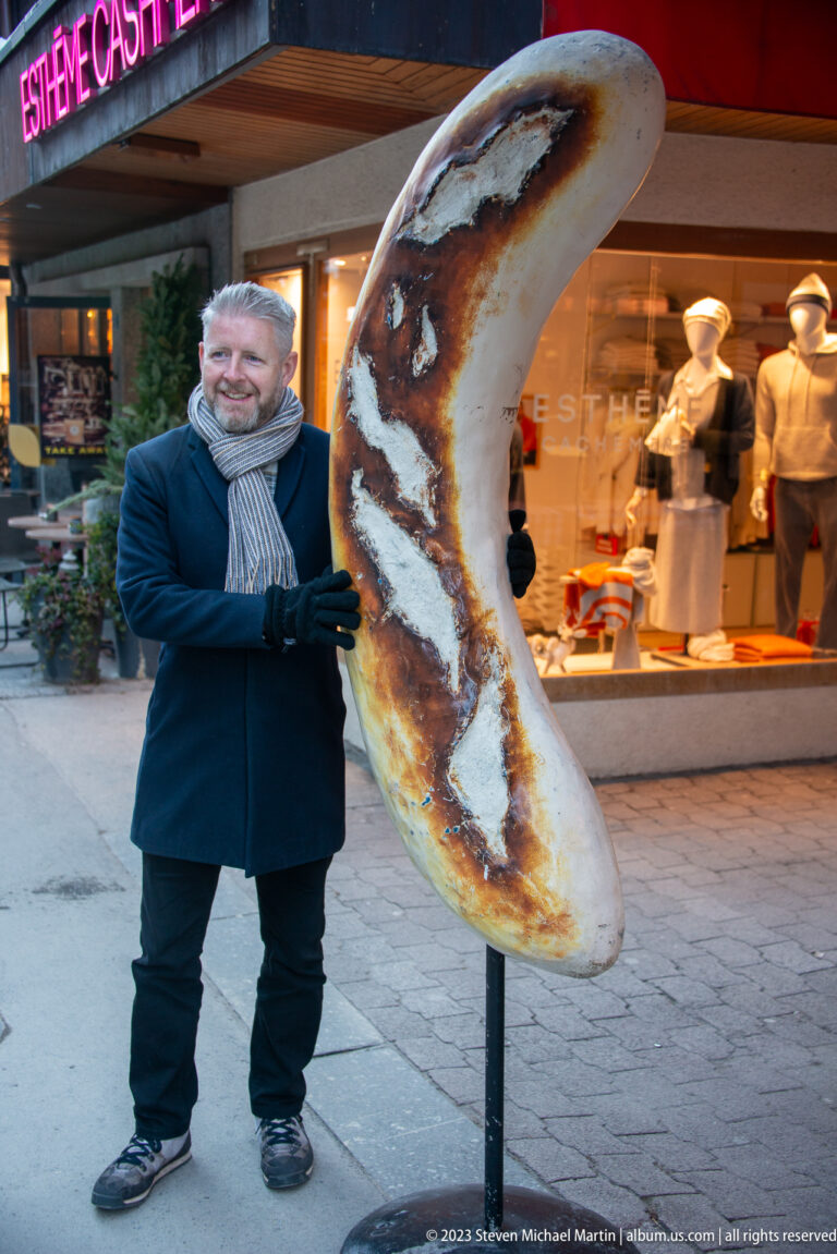 Man in winter coat and scarf posing beside an oversized crescent-shaped pastry sculpture outside a shop.