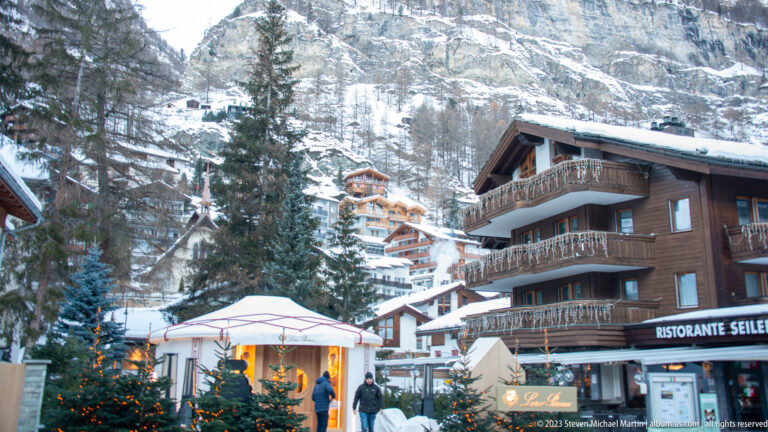 Chalet-style hotel, decorated Christmas tree, and white canopy below steep snow-covered cliffs in Zermatt.