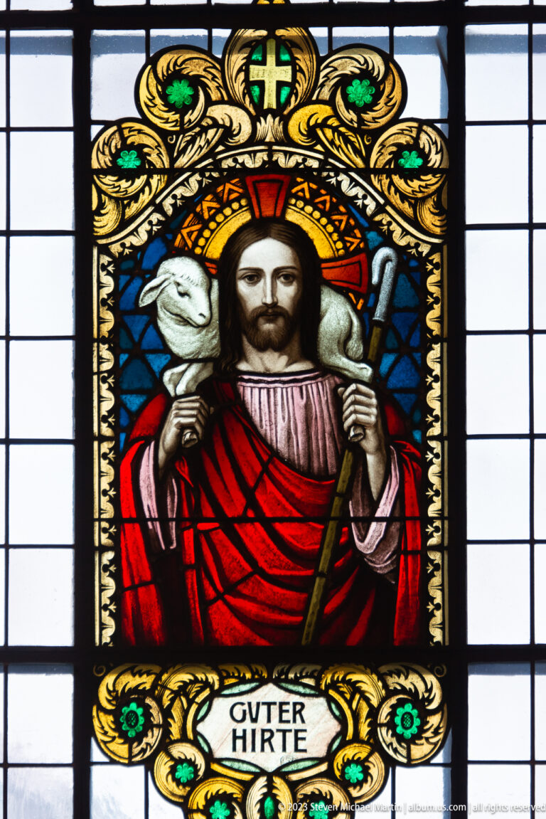 Stained-glass Christ as the Good Shepherd carrying a lamb and staff above the inscription GUTER HIRTE.