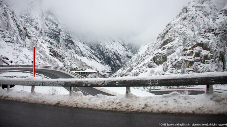 Snowy mountain gorge with curving roads and bridges between steep rock walls under low cloud.