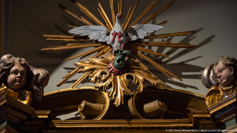 White Holy Spirit dove before gilded rays above an ornate altar, flanked by carved cherub heads.