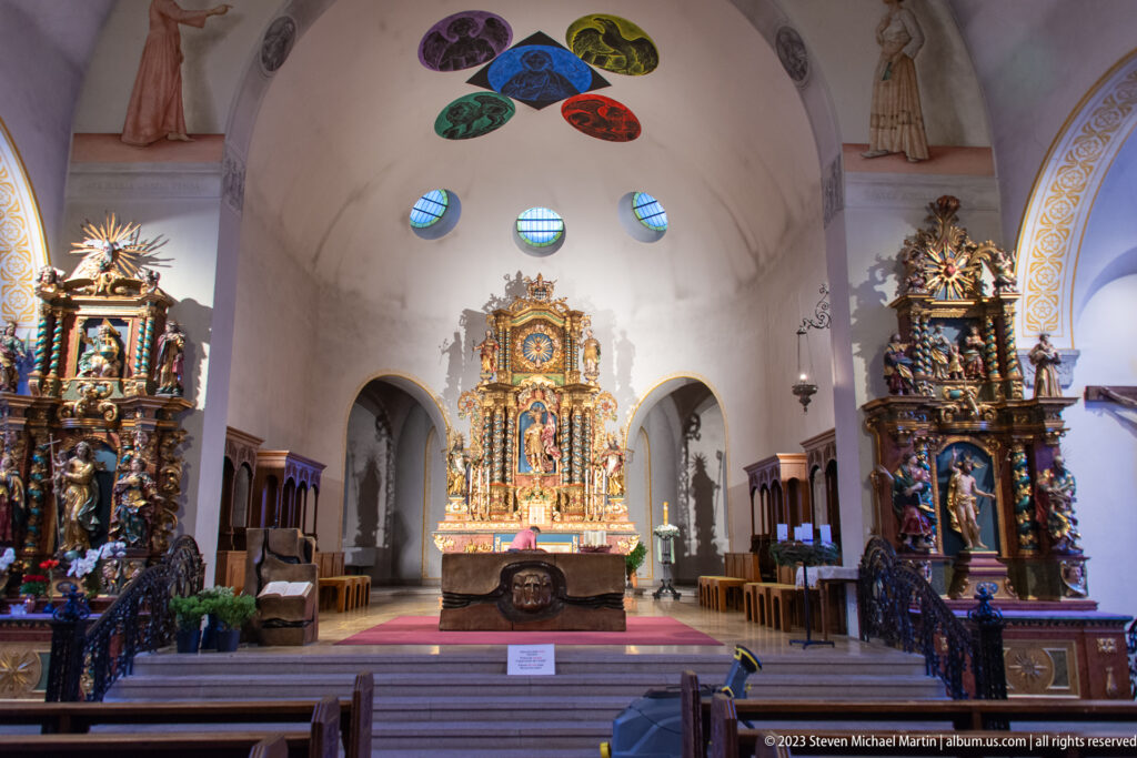 Church interior with gilded central altar, richly carved side altars, vaulted white walls, and small stained-glass windows.