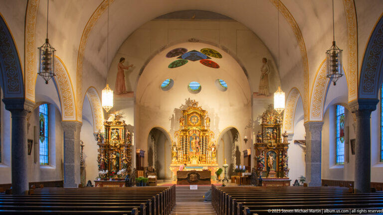 Symmetrical nave view with wooden pews leading to a gilded high altar and two carved side altars.