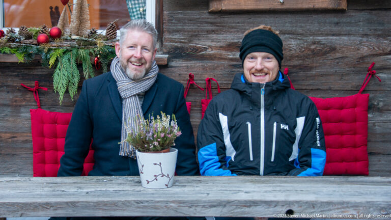 Two smiling men in winter clothing seated on a red-cushioned bench against a timber wall.