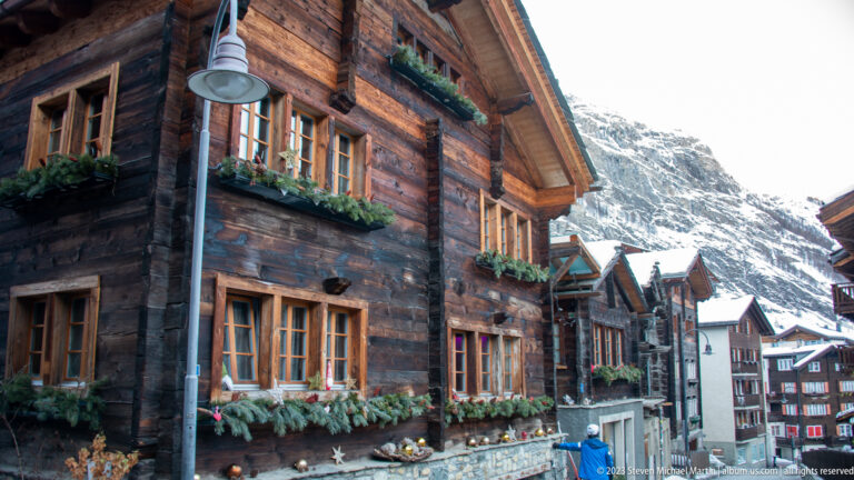 Dark weathered timber chalet with many small-paned windows, evergreen garlands, and flower boxes.