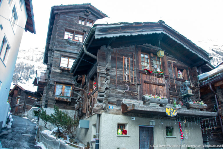 Weathered timber house with hanging rustic decorations, garlands, flower boxes, and a pale masonry ground floor.
