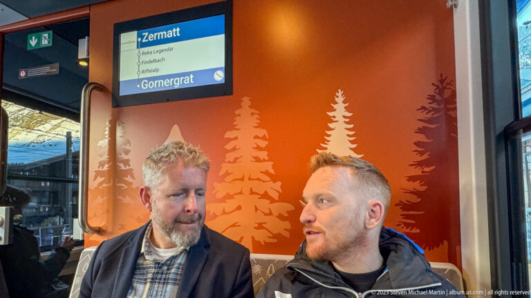 Two men seated inside a mountain railway carriage beneath a digital display showing Gornergrat.