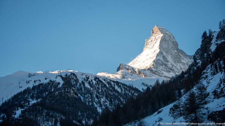 Matterhorn glowing in sunlight above shadowed snowy forested slopes under a clear blue sky.