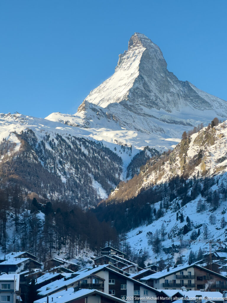 Broad view of the Matterhorn above snowy slopes, forest, and scattered buildings near Zermatt.