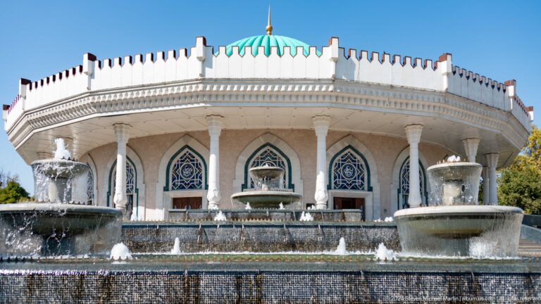 White colonnaded museum facade with pointed arches, blue decorative windows, turquoise dome, and tiered fountains under a clear sky.