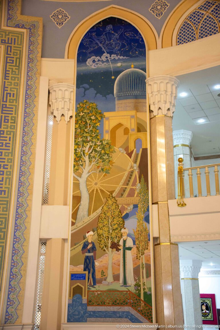 Vertical painted panel with a large spoked wheel, leafy tree, tall tower, small figures, and blue geometric border.