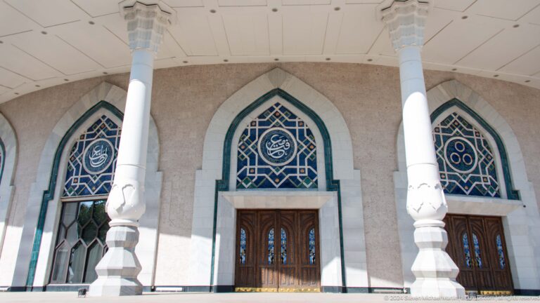 Pointed-arch museum entrance with blue geometric glazing, circular calligraphic medallion, carved wooden doors, and white columns.