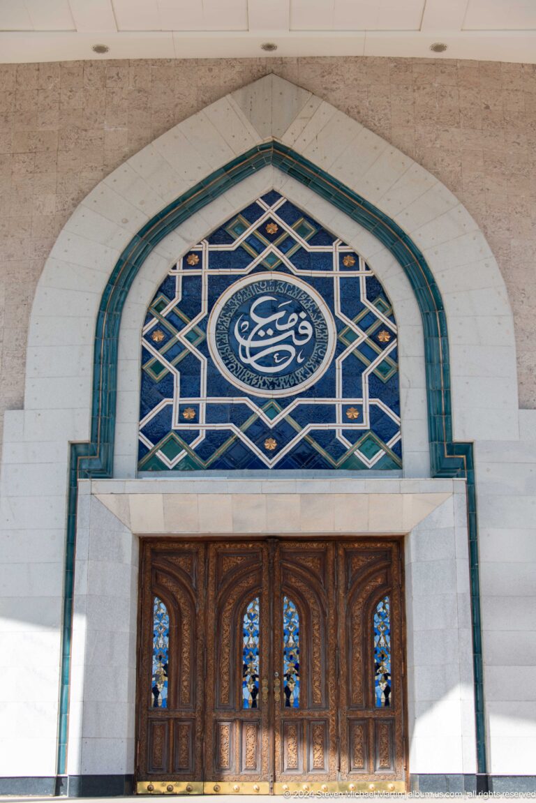 Close view of a pointed-arch blue geometric window with a circular calligraphic medallion above carved wooden doors.
