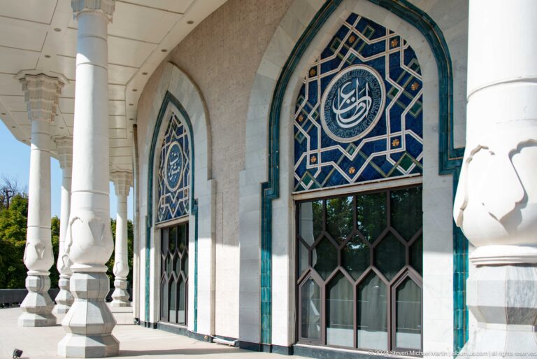 Oblique view along a white museum arcade with repeated columns, pointed arches, blue geometric windows, and dark entrance doors.
