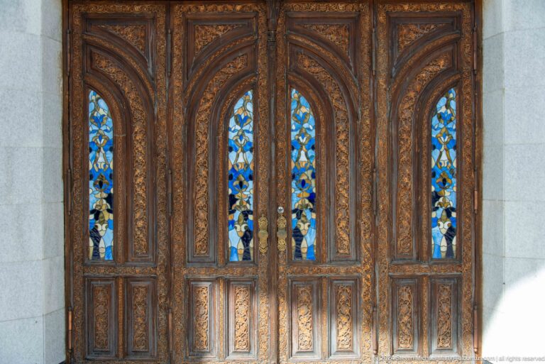 Ornately carved dark wooden double doors with narrow pointed panels of blue and yellow decorative glass.