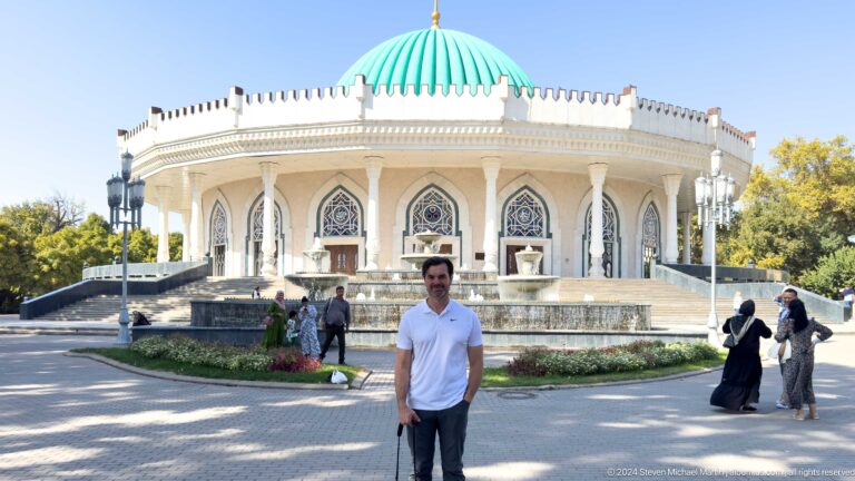 Man in white shirt before turquoise dome and arcaded facade.