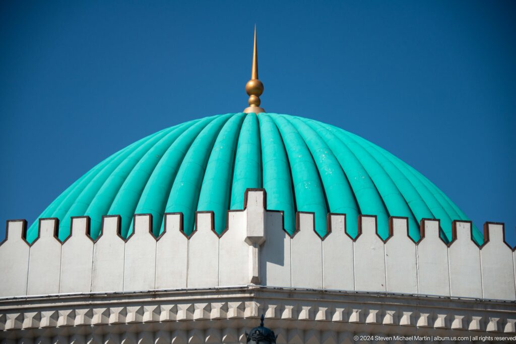 Turquoise ribbed museum dome with a gold finial above a white crenellated parapet against a deep blue sky.