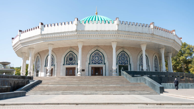 Broad stone steps leading to three pointed-arch museum entrances beneath white columns and a turquoise dome.