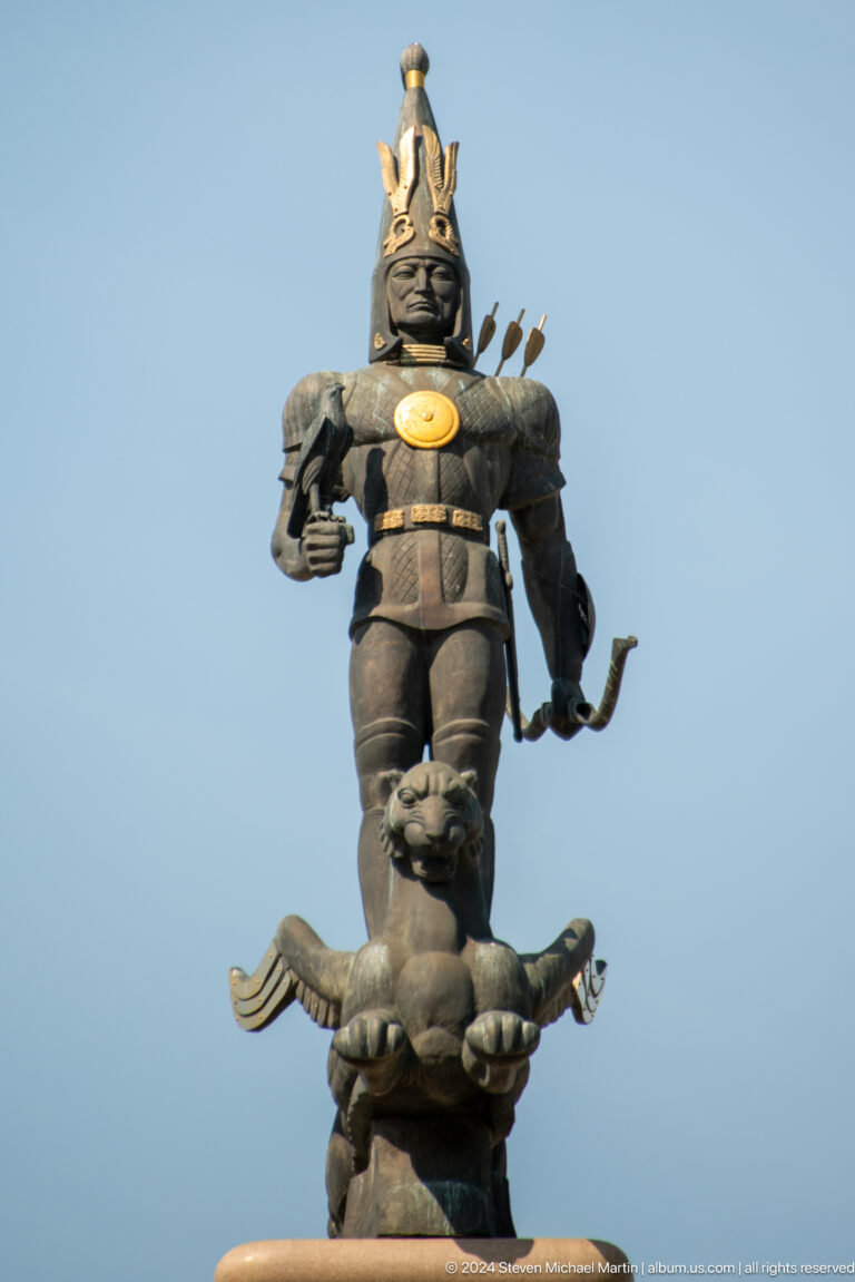 Frontal view of the Golden Warrior monument in Almaty, showing the bronze armored figure standing atop a sculptural base with a feline head.