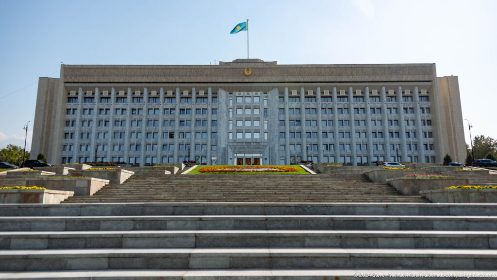 Wide stone steps leading up to the large, rectangular Almaty city administration building with rows of windows and flagpoles in front.