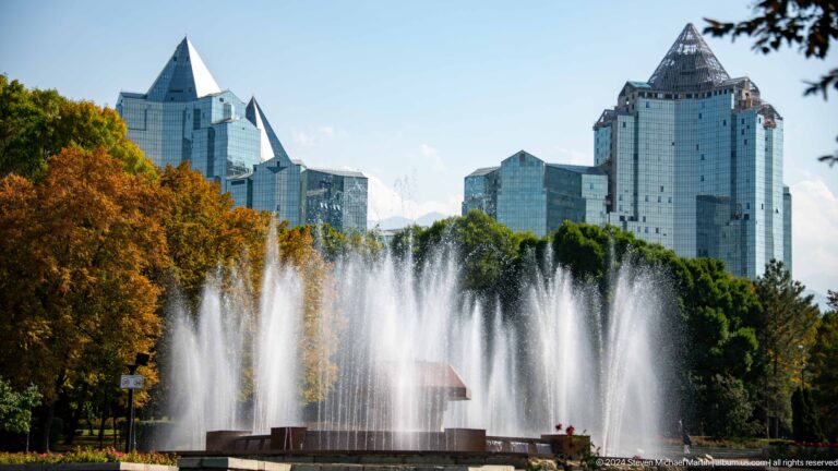 Large Republic Square fountains in Almaty spraying water in front of modern high-rise buildings under a hazy sky.