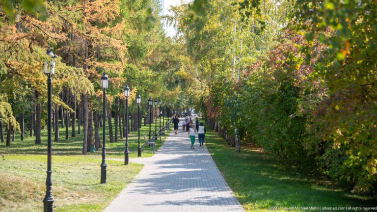 Long straight pedestrian path lined with trees in an Almaty urban park, with people walking along the promenade.