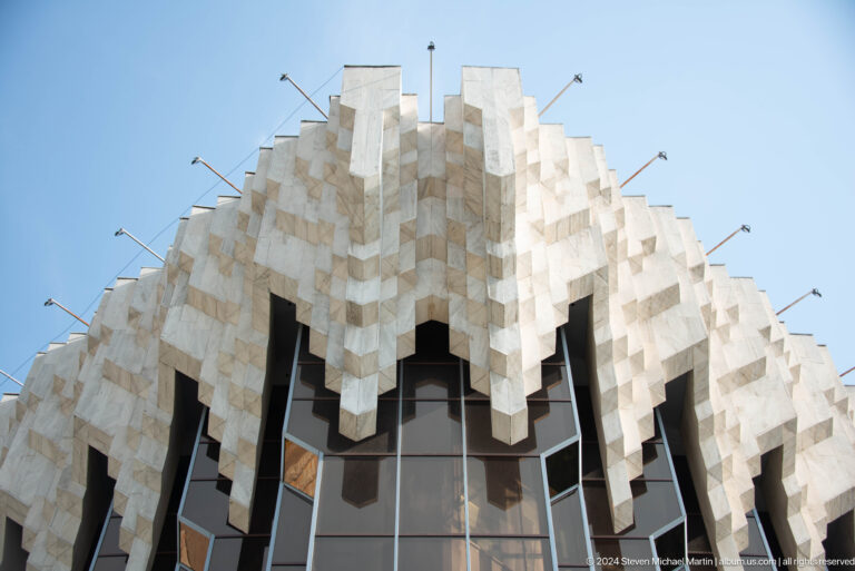 Low-angle view of the Palace of the Republic facade in Almaty, showing a faceted white concrete canopy above dark reflective glazing.
