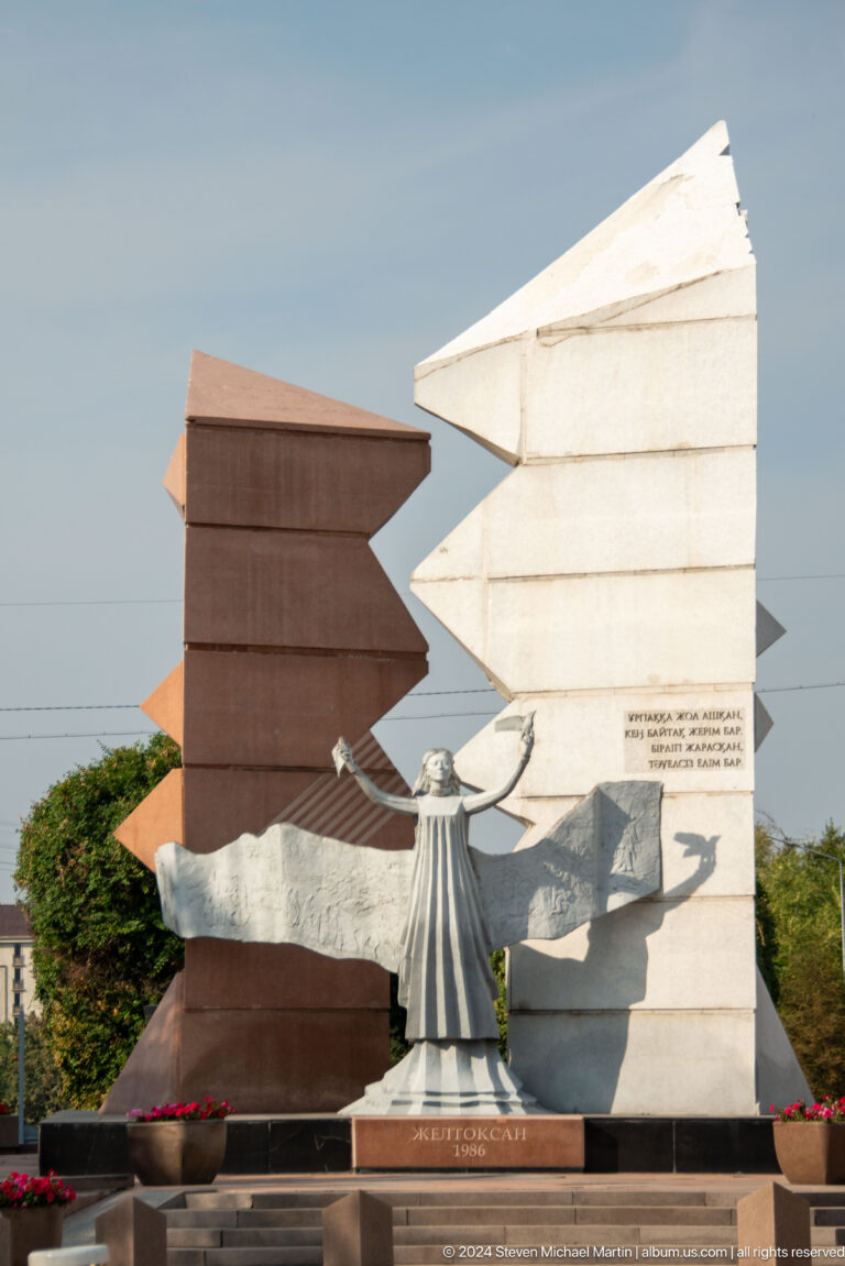 Abstract public memorial in Almaty with a crescent-like metal form inset into a stone wall in a landscaped park.