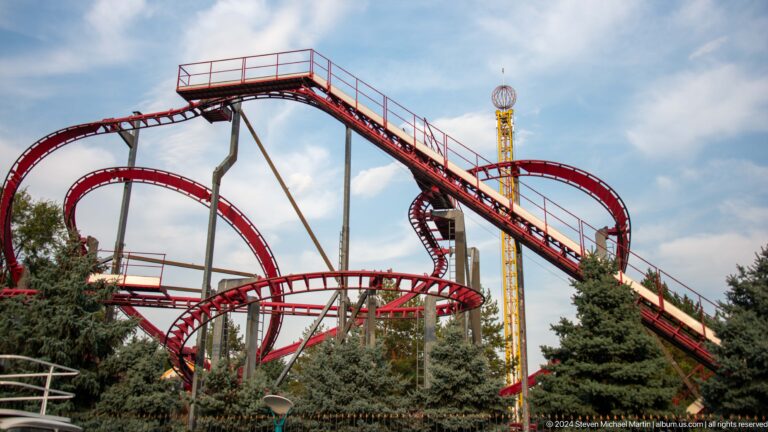 Red roller coaster track curving through trees in an outdoor amusement park.