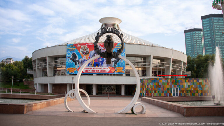 Exterior of the Kazakh State Circus in Almaty with a large white circular sculpture in front of the arena facade.