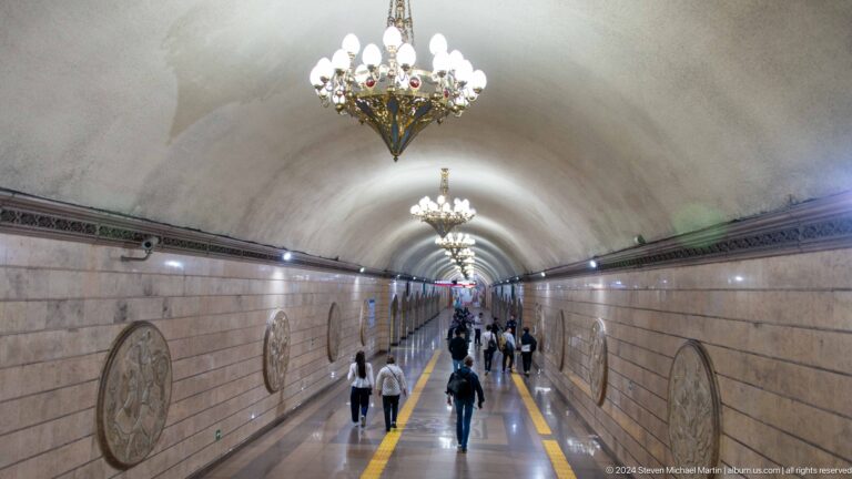 Ornate Almaty Metro station hall with chandeliers hanging over a wide stone platform and tracks.