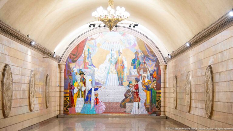 Wide view of an Almaty metro hall with a vaulted ceiling, ending in a large colorful Kazakh historical mosaic mural.