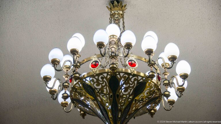 Low-angle close-up of an ornate multi-globe chandelier in a metro station, with white globes on dark metal scrollwork and red gem-like details.