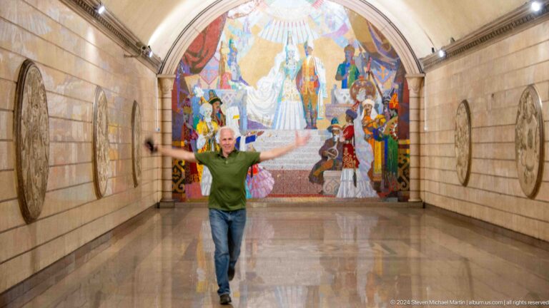 Visitor in a green shirt standing in front of a large historical mosaic on the arched end wall of a marble-lined metro station interior.