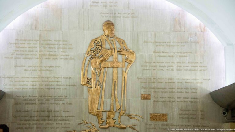 Bas-relief of a Kazakh man in traditional dress on a marble wall inside an Almaty Metro station, surrounded by inscriptions.