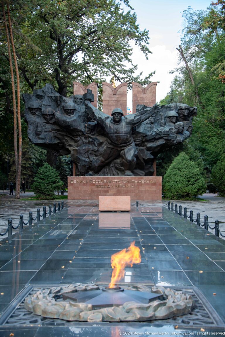Straight-on view along the memorial plaza in Almaty aligned toward the bronze Panfilov war memorial sculpture group.