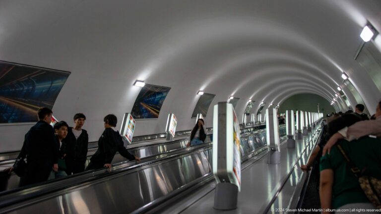 People standing and riding on parallel escalators in a brightly lit arched metro tunnel in Almaty.