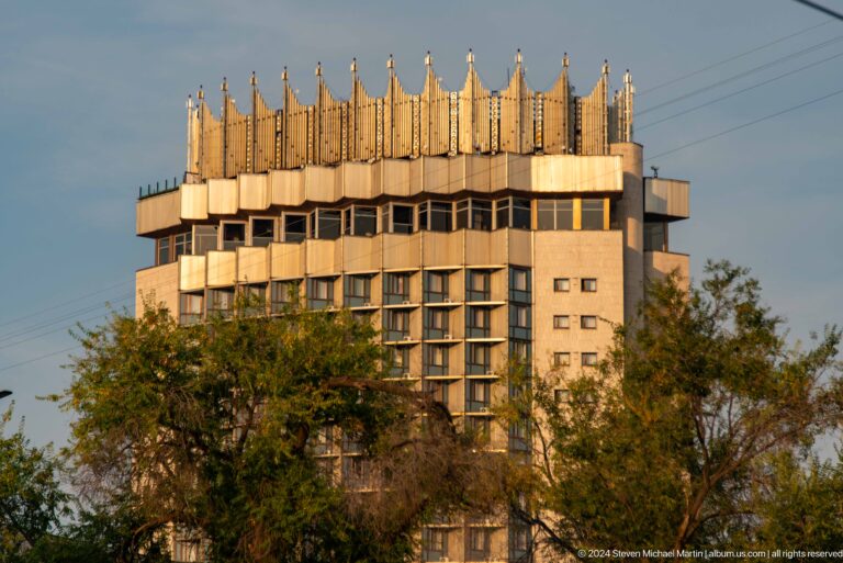 Soviet-era high-rise tower in Almaty with a faceted façade and ornate metal crown rising above surrounding trees.