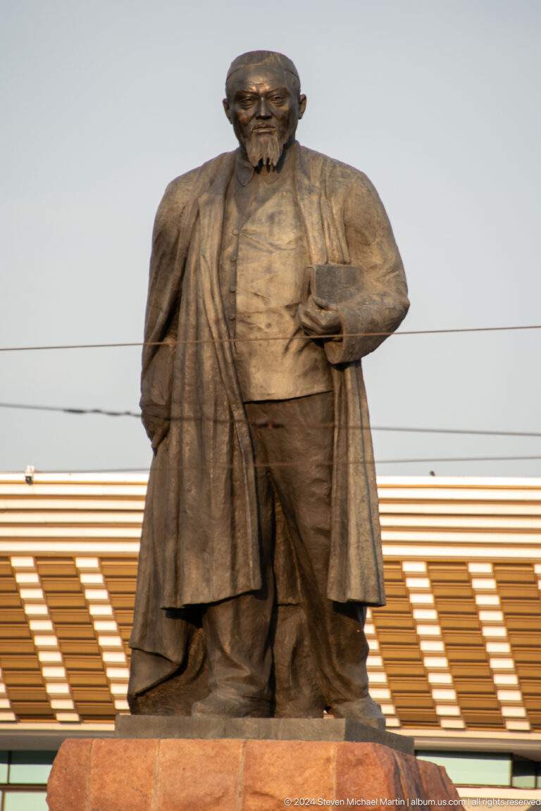 Bronze statue of a bearded man with a book on a stone pedestal in front of a modern building in Almaty.