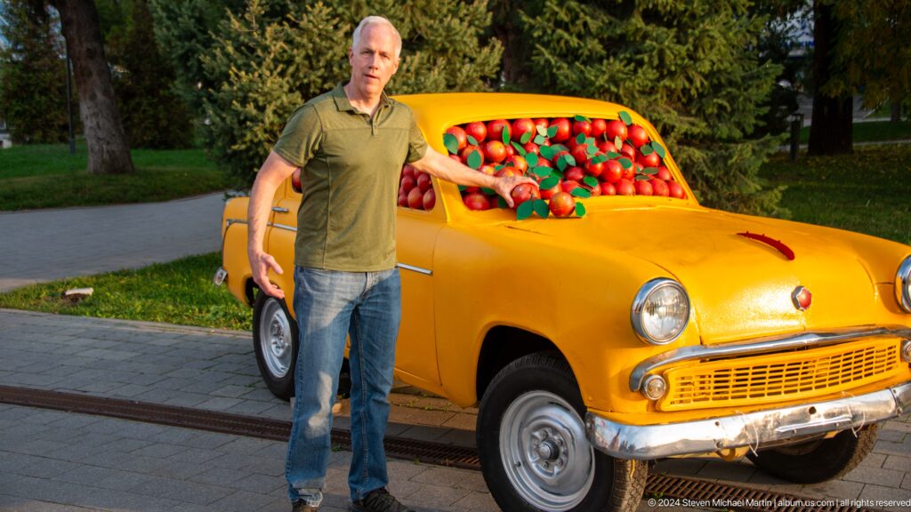 Man standing by a yellow vintage-style car sculpture whose interior and windshield are filled with bright red apples in a park in Almaty.