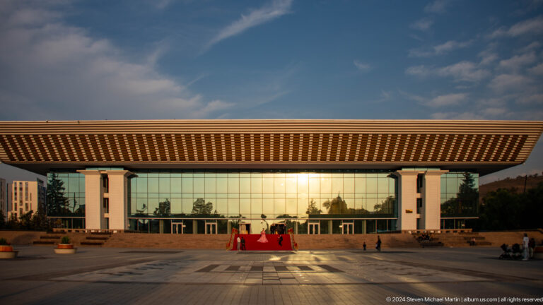 Modern glass building with a broad cantilevered roof in Almaty, with a couple standing on a red carpet in the center of the plaza at sunset.