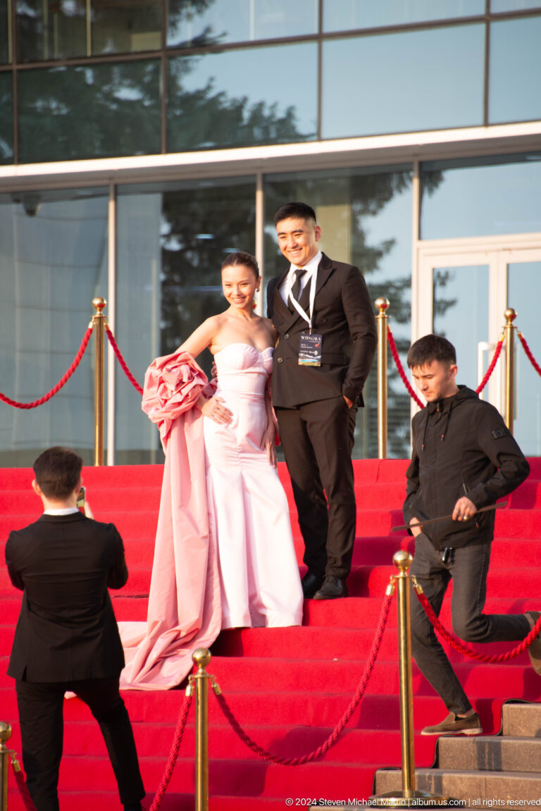 Formally dressed couple posing for photos on a red carpeted staircase outside a glass-fronted building in Almaty.