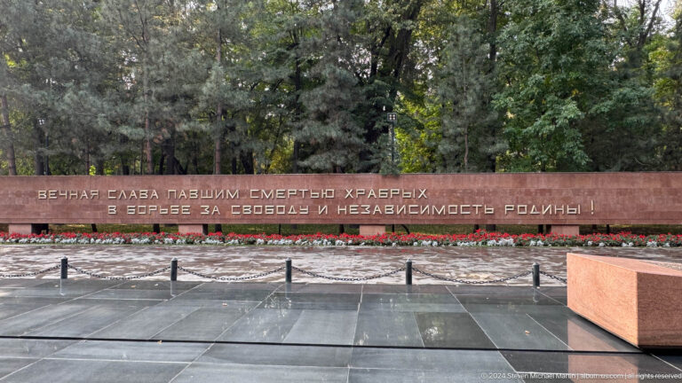 Wide view of a long red-brown memorial inscription wall in Panfilov Park, Almaty, with trees behind and a paved forecourt in front.