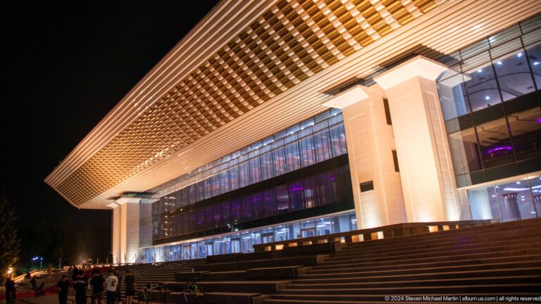 Night view of broad steps leading to a glass-fronted modern building entrance beneath a deep illuminated soffit in Almaty, Kazakhstan.
