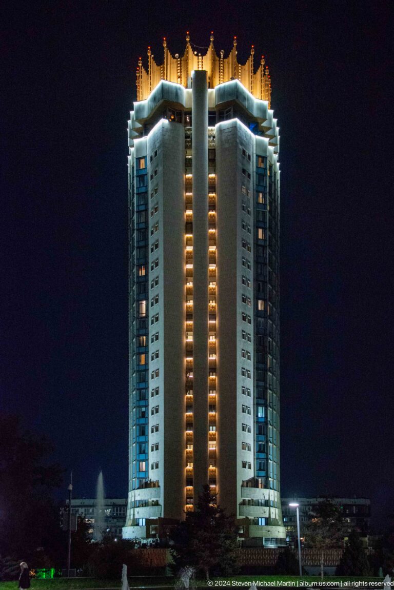 Night view of a tall illuminated high-rise hotel tower in Almaty with vertical bands of lit windows against a dark sky.