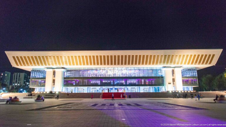 Wide nighttime view of an illuminated hotel complex in Almaty with a bright horizontal canopy across the facade.