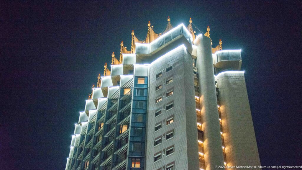 Night view of an illuminated high-rise tower in Almaty, Kazakhstan, with stepped balconies and decorative lighting against a dark sky.